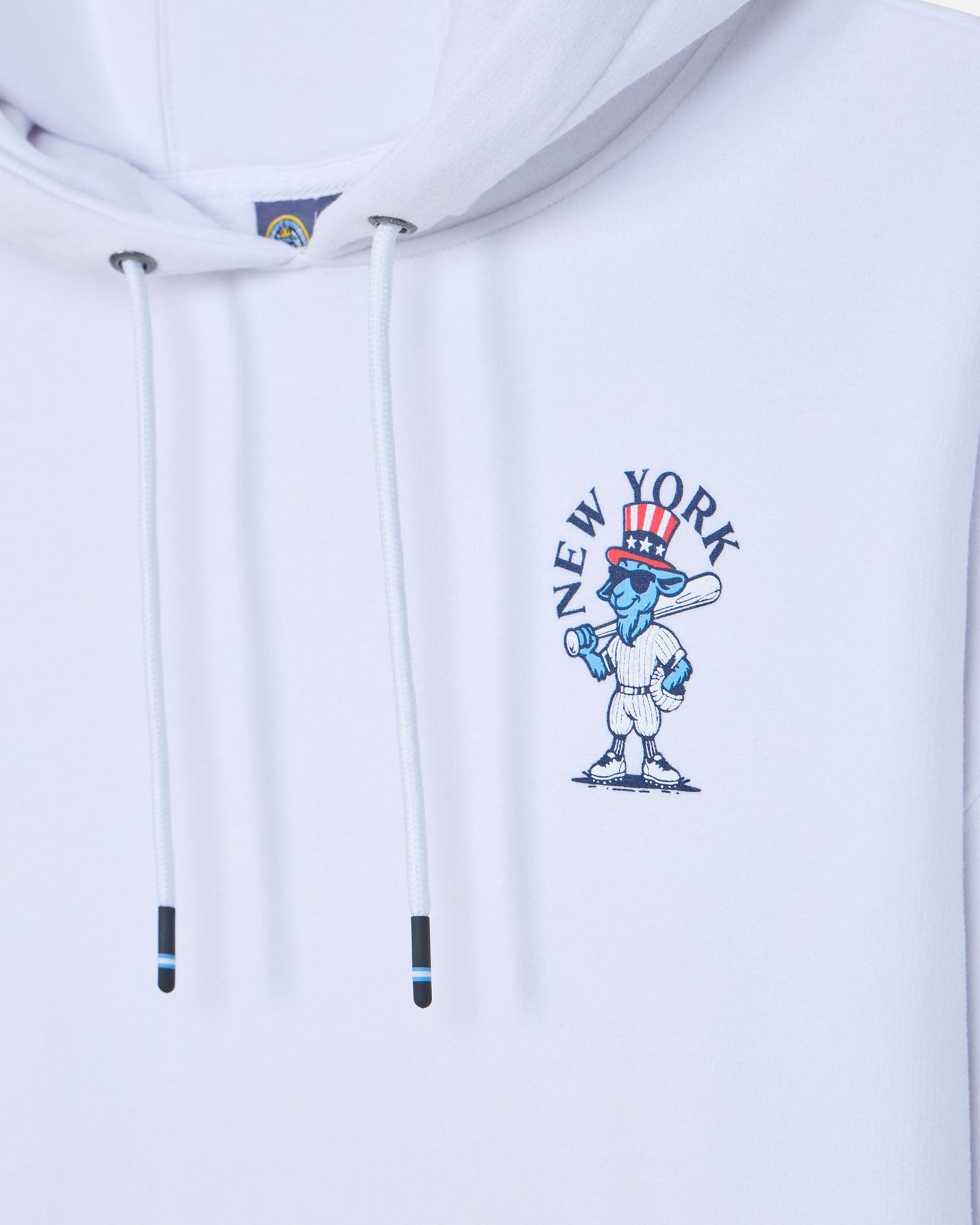 Close-up of a white pullover hoodie with white drawstrings and a blue, red, and white New York City graphic on the chest.#color_white