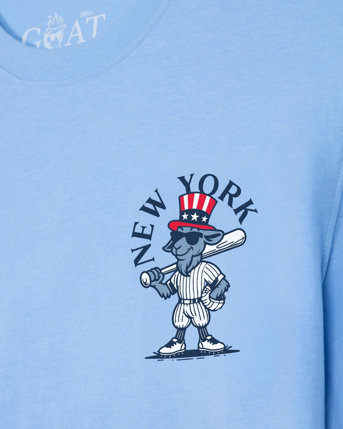 A close-up of a light blue t-shirt featuring a cartoon graphic of a gray goat baseball player wearing a red, white, and blue Uncle Sam hat, with the text 'NEW YORK' arched above him. A white 'GOAT' brand logo is visible on the inner neck collar.#color_carolina-blue