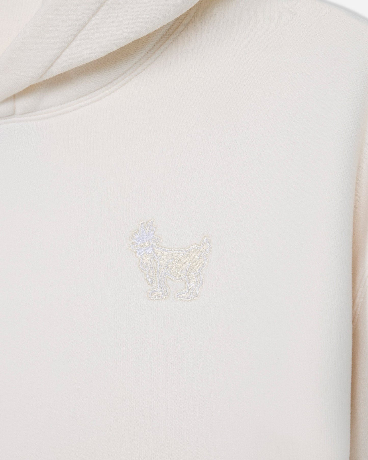 Close-up of a cream-colored hoodie featuring a small, tonal embroidered goat logo on the chest.#color_bone