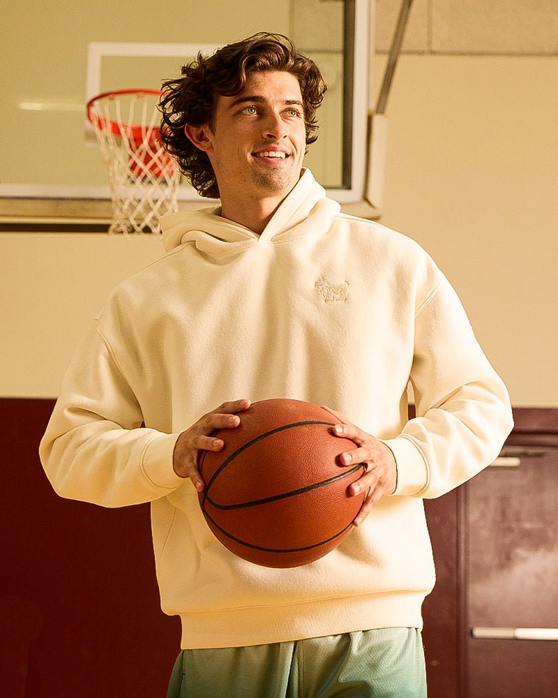 A cream-colored long-sleeve hoodie with a small tonal embroidered logo on the left chest, on an adult male model.#color_bone