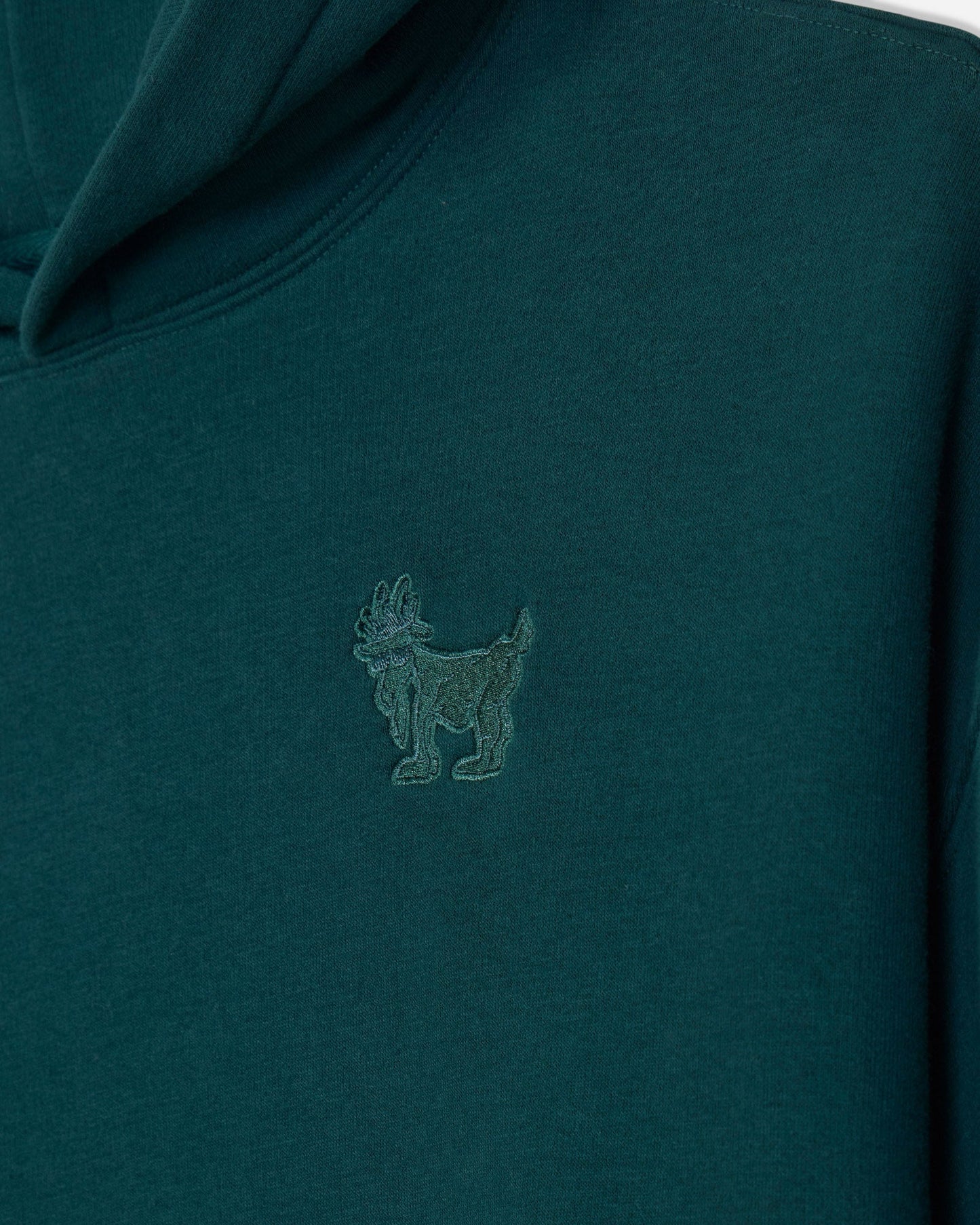 Close-up of a dark teal hooded sweatshirt featuring a tonal embroidered goat logo on the left chest.#color_sage