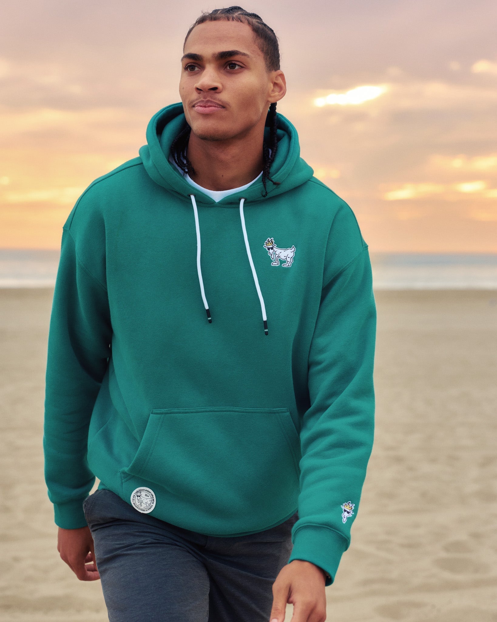 Man wearing a teal hoodie with a logo on a beach at sunset#color_teal-blue