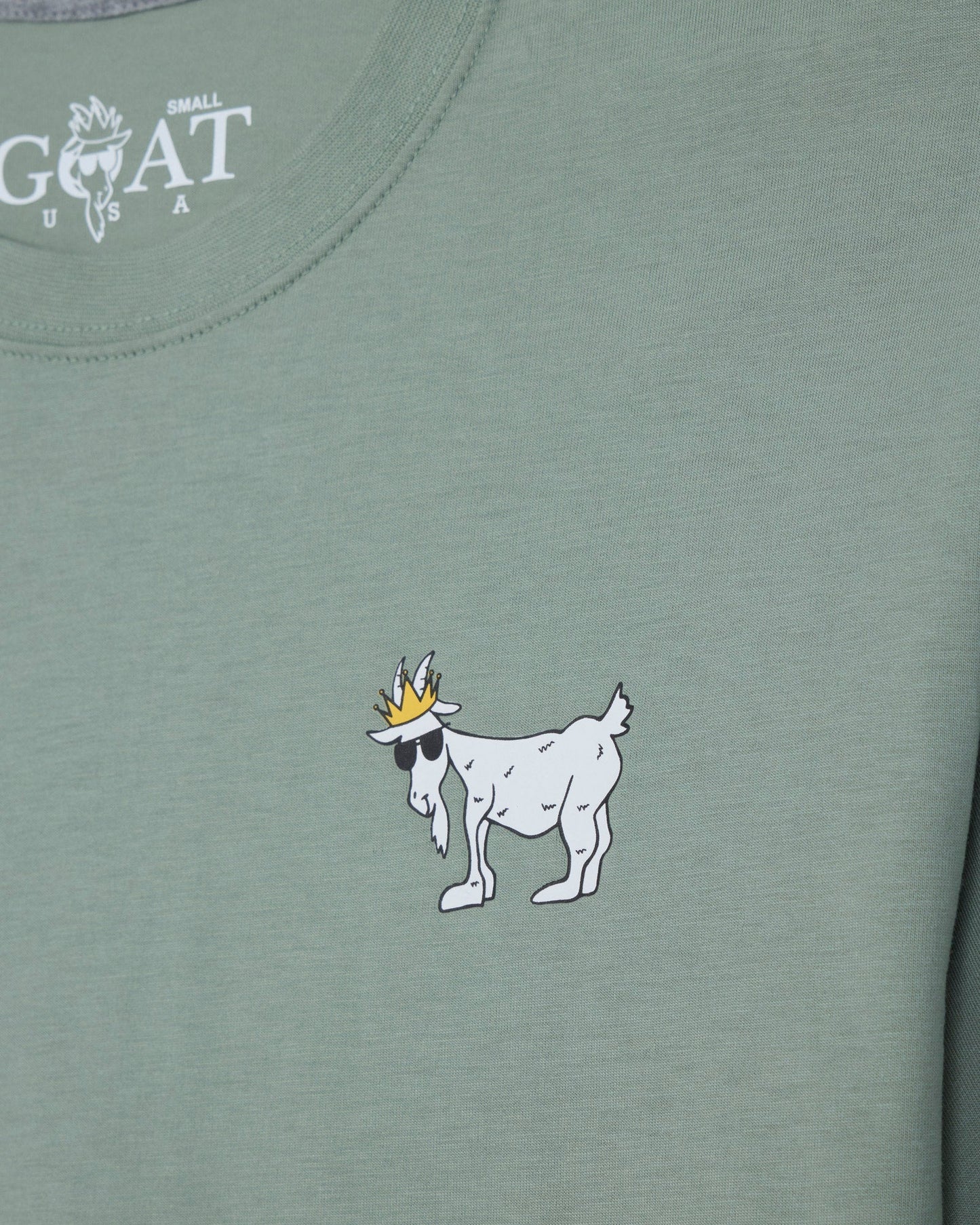 Close-up of a sage green t-shirt featuring a white cartoon goat wearing a gold crown on the chest, with the white 'GOAT' brand logo printed inside the collar.#color_steeped-sage