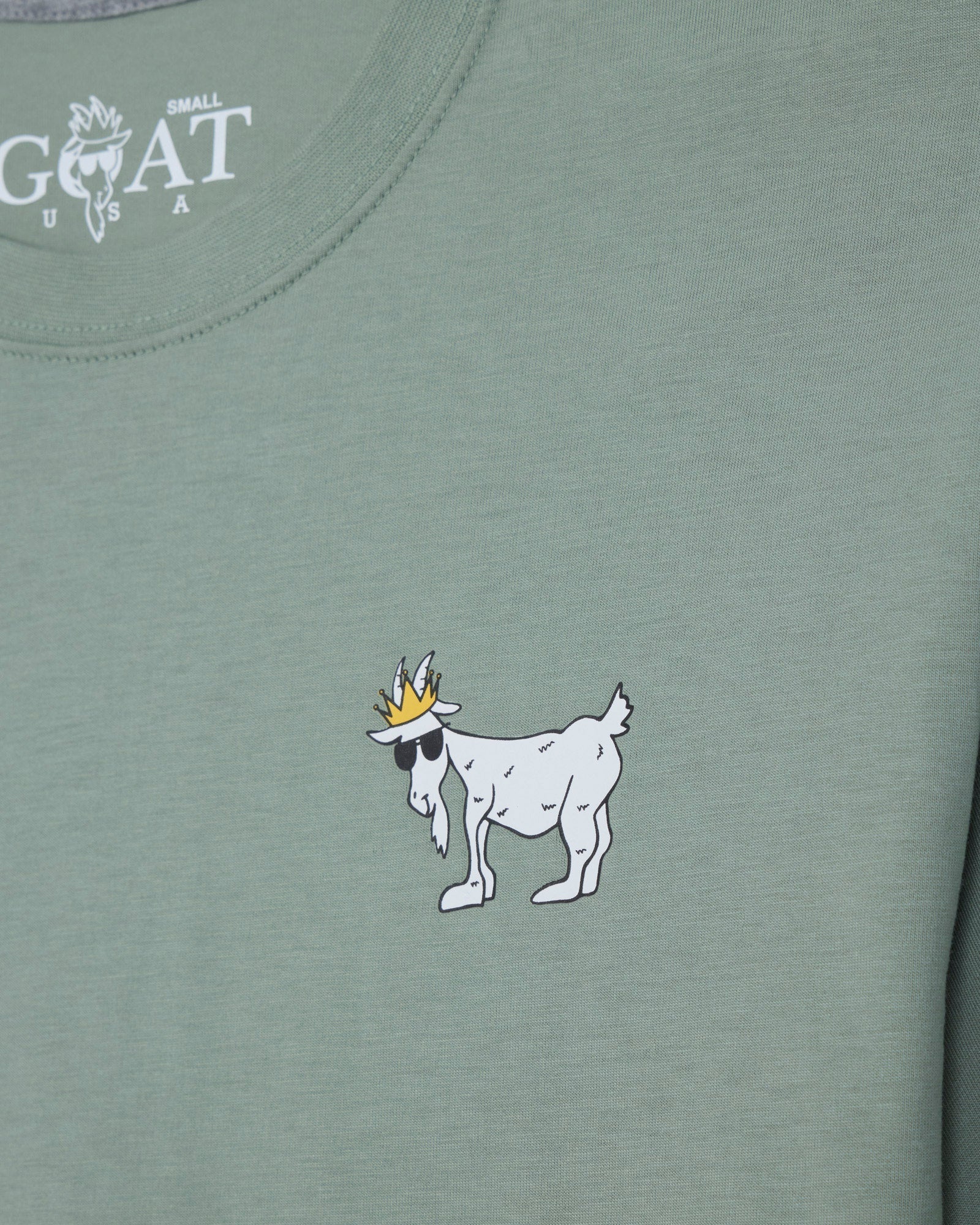 Close-up of a sage green t-shirt featuring a white cartoon goat wearing a gold crown on the chest, with the white 'GOAT' brand logo printed inside the collar.#color_steeped-sage