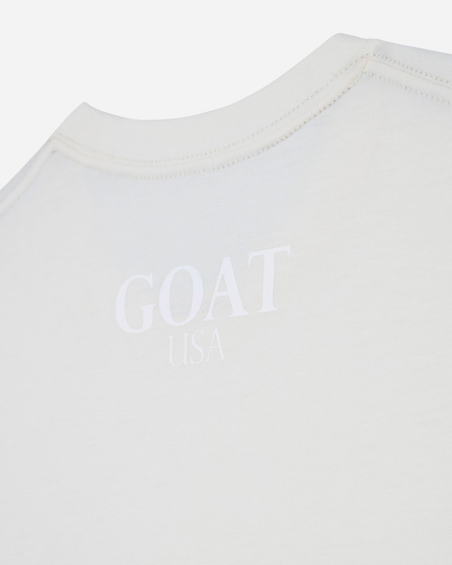 Close-up of a white crewneck t-shirt with a tonal, embossed 'GOAT USA' logo printed on the chest.#color_bone
