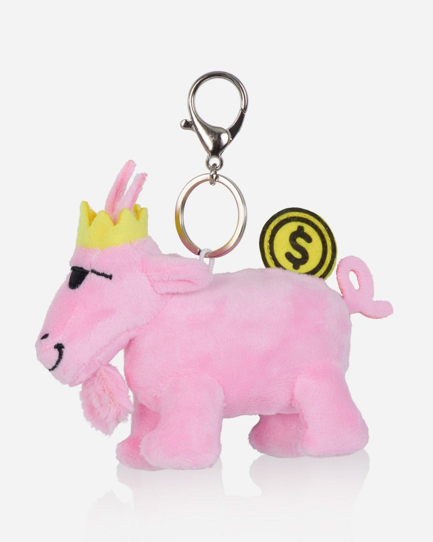 A pink plush goat keychain charm with a yellow crown, black embroidered eyes and mouth, a silver metal clasp, and a yellow coin-shaped tag with a dollar sign.