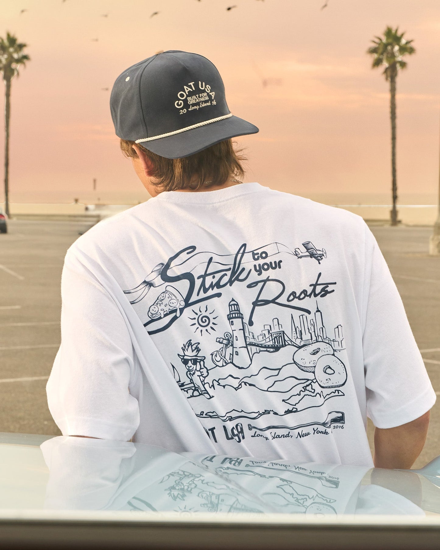 Person wearing a white t-shirt with a graphic design and a cap, sitting by a beach with palm trees.