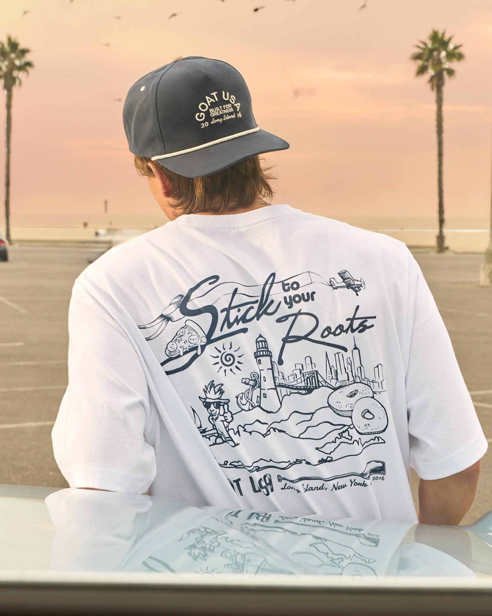 Person wearing a white t-shirt with a graphic design and a cap, sitting by a beach with palm trees.