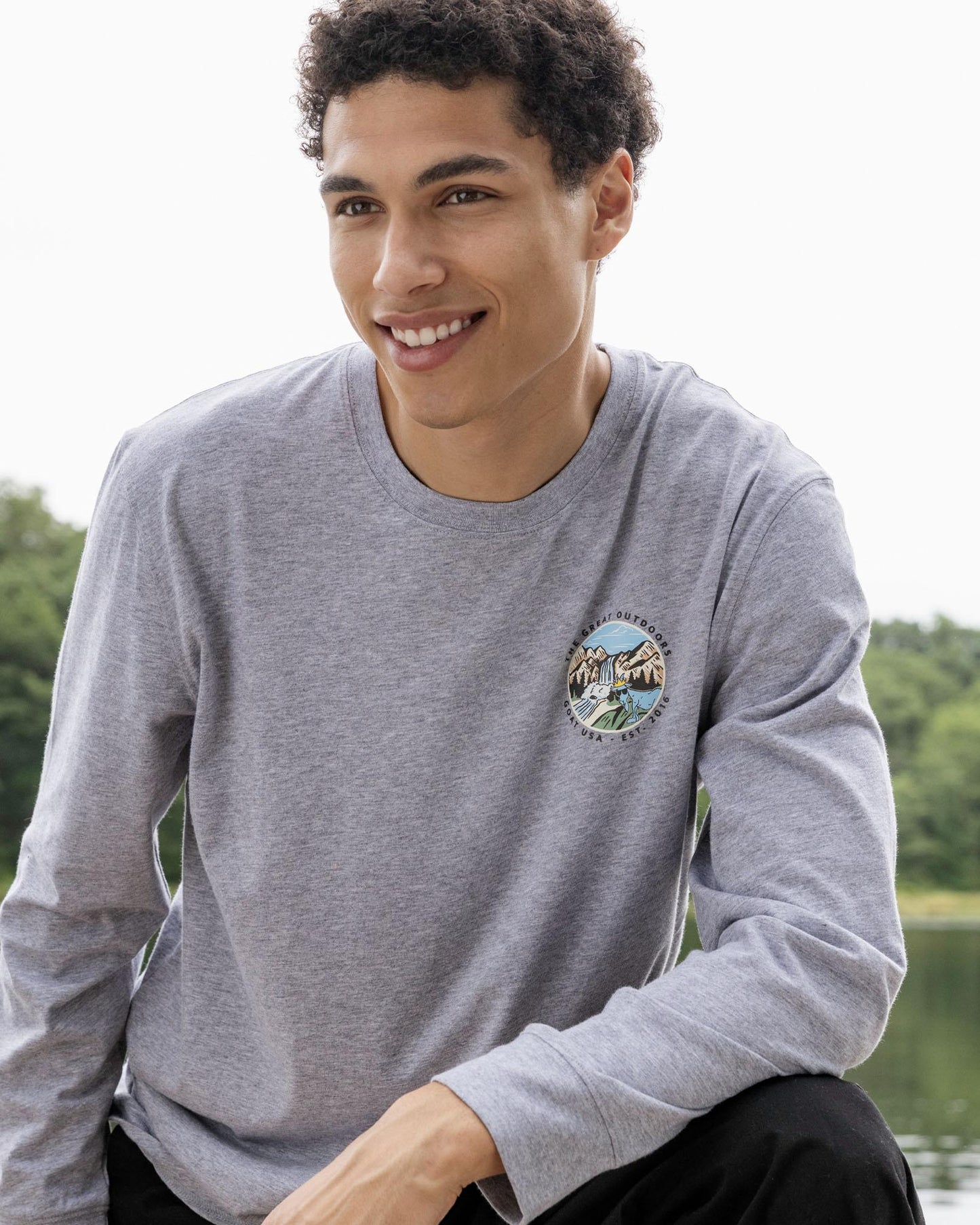 Man wearing a gray long-sleeve shirt with a circular design, sitting by a body of water.