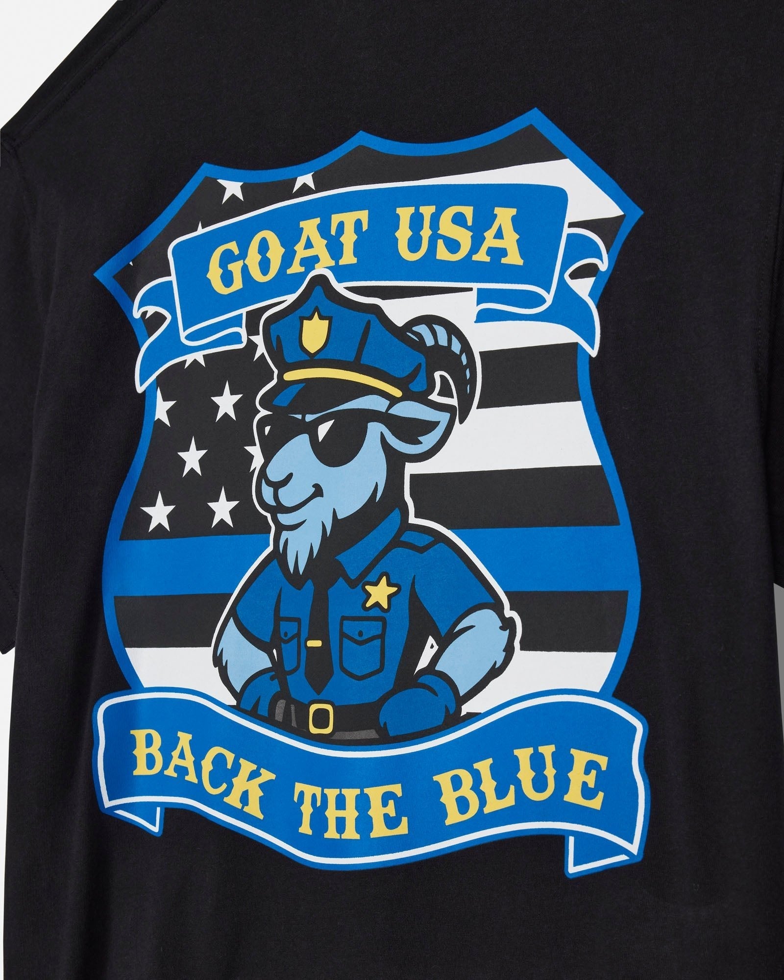 Close-up of a black T-shirt featuring a large graphic print of a cartoon goat in a police uniform with the text 'GOAT USA' and 'BACK THE BLUE' on blue banners, set against a black and white striped shield design.