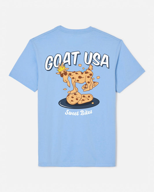 A light blue short-sleeve t-shirt shown from the back, featuring a cartoon graphic of a goat wearing a crown and standing on chocolate chip cookies, with the text 'GOAT USA' and 'Sweet Bites'.