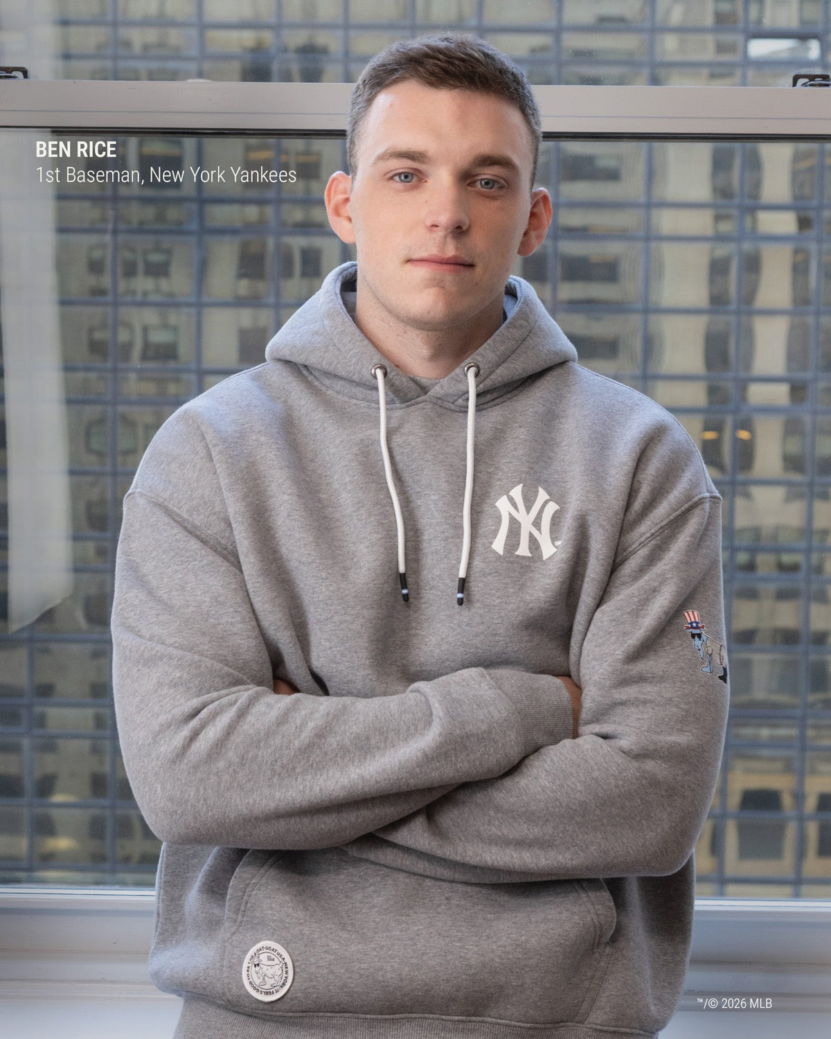 A man wearing a gray New York Yankees hoodie stands with arms crossed in front of a window showing city buildings. The hoodie has a white NY logo on the chest, white drawstrings, and a small sleeve patch.