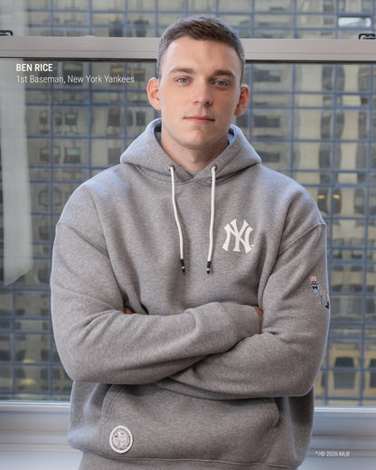 A man wearing a gray New York Yankees hoodie stands with arms crossed in front of a window showing city buildings. The hoodie has a white NY logo on the chest, white drawstrings, and a small sleeve patch.