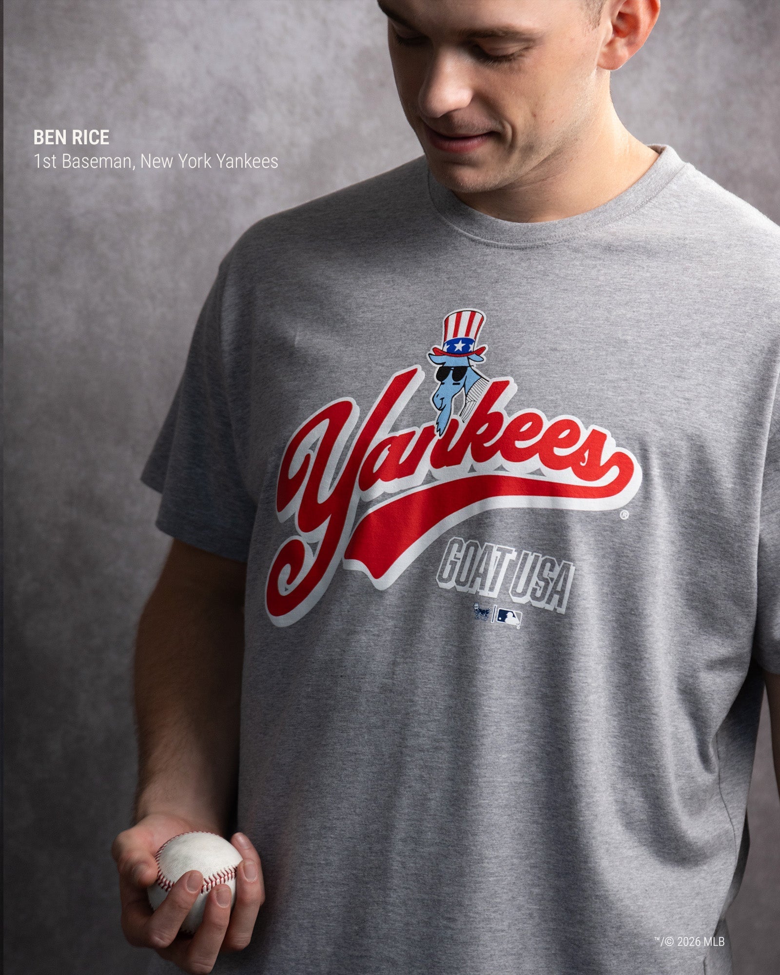 A man wearing a heather gray New York Yankees graphic T-shirt, holding a baseball. The shirt features a red and white 'Yankees' script logo with a blue character wearing an Uncle Sam hat, and 'GOAT USA' printed below.
