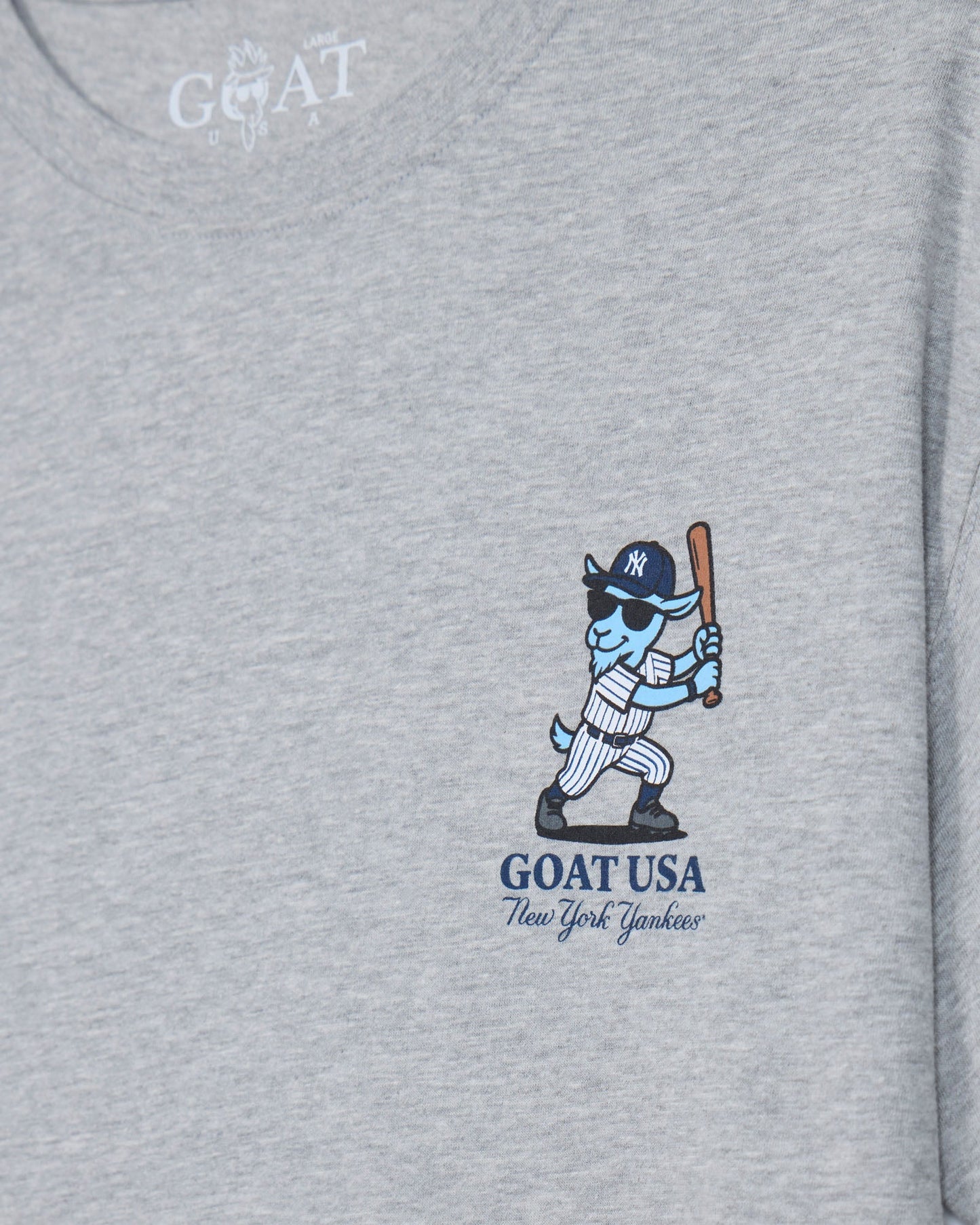 Close-up of a heather gray t-shirt featuring a graphic of a blue and white goat baseball player holding a bat, with 'GOAT USA New York Yankees' printed below.#color_gray
