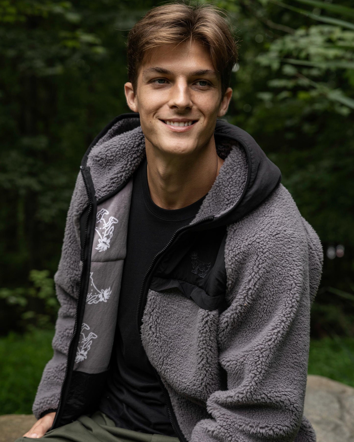 Man wearing a gray fleece jacket with a black shirt underneath, sitting outdoors with greenery in the background.#color_gray