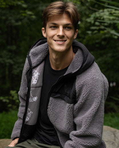Man wearing a gray fleece jacket with a black shirt underneath, sitting outdoors with greenery in the background.#color_gray